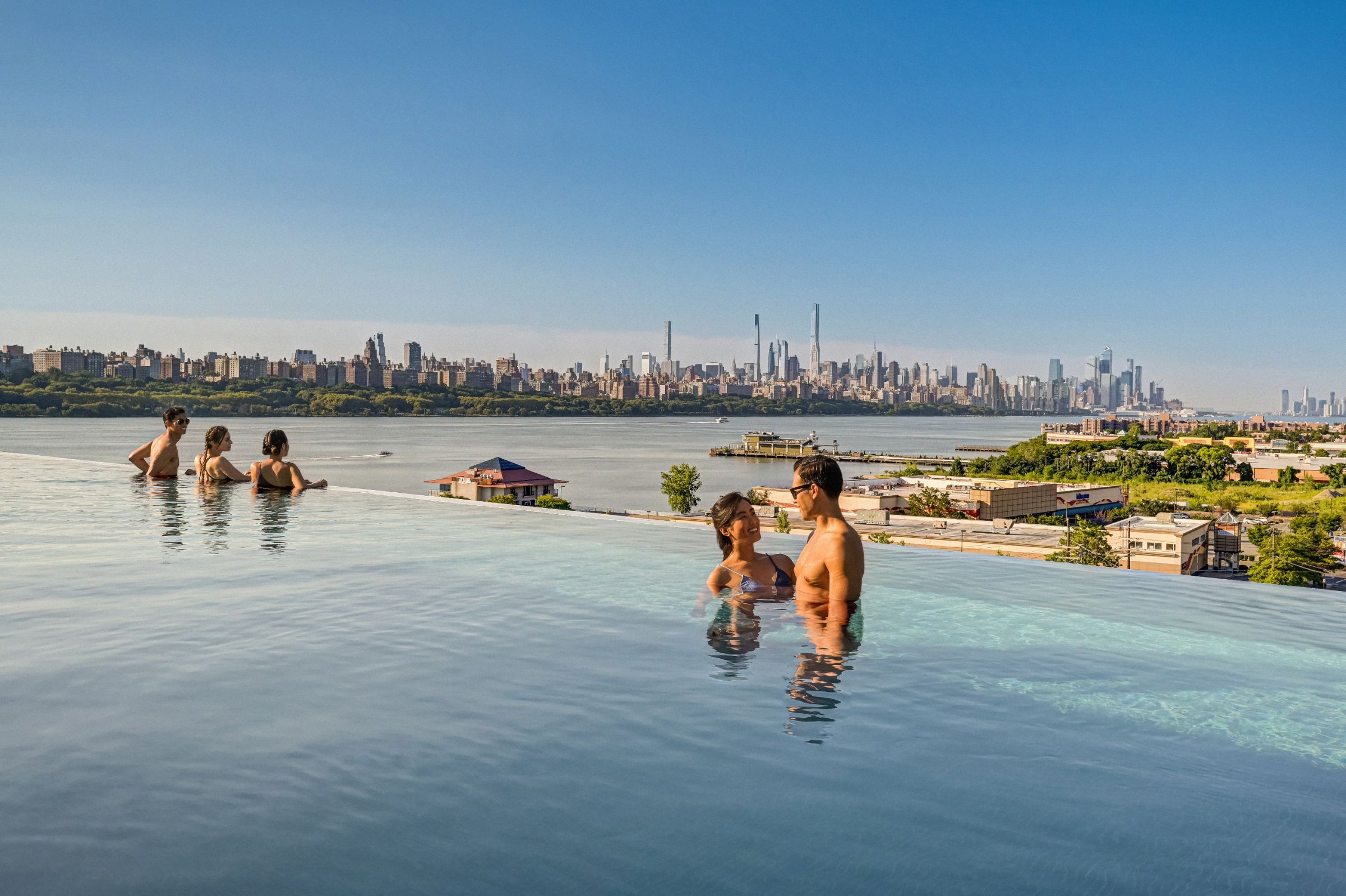 A smiling couple & 3 friends enjoy NYC skyline from SoJo spa club's rooftop infinity pool on a bright, sunny day