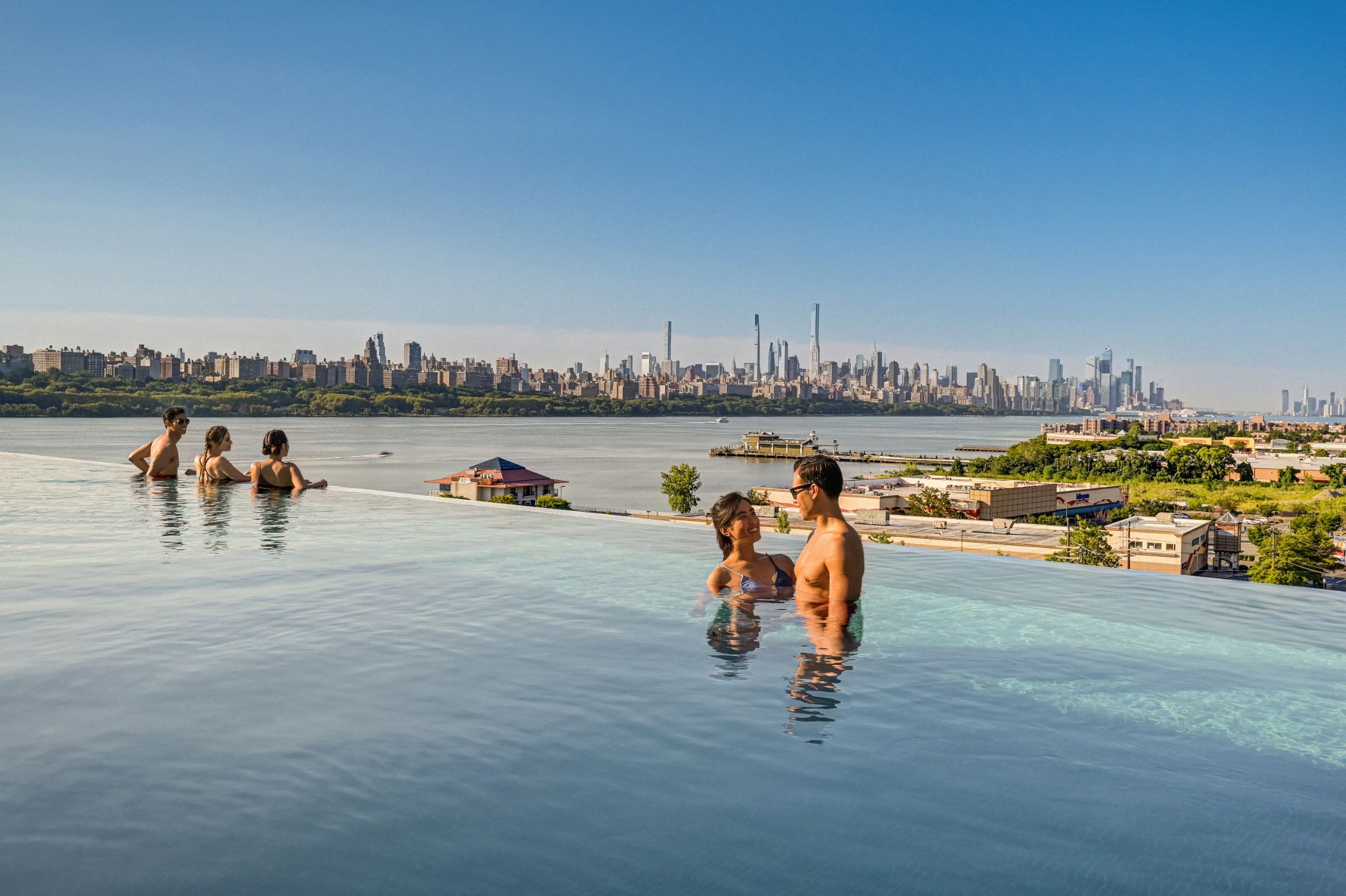 A smiling couple & 3 friends enjoy NYC skyline from SoJo Spa Club's rooftop infinity pool on a bright, sunny day