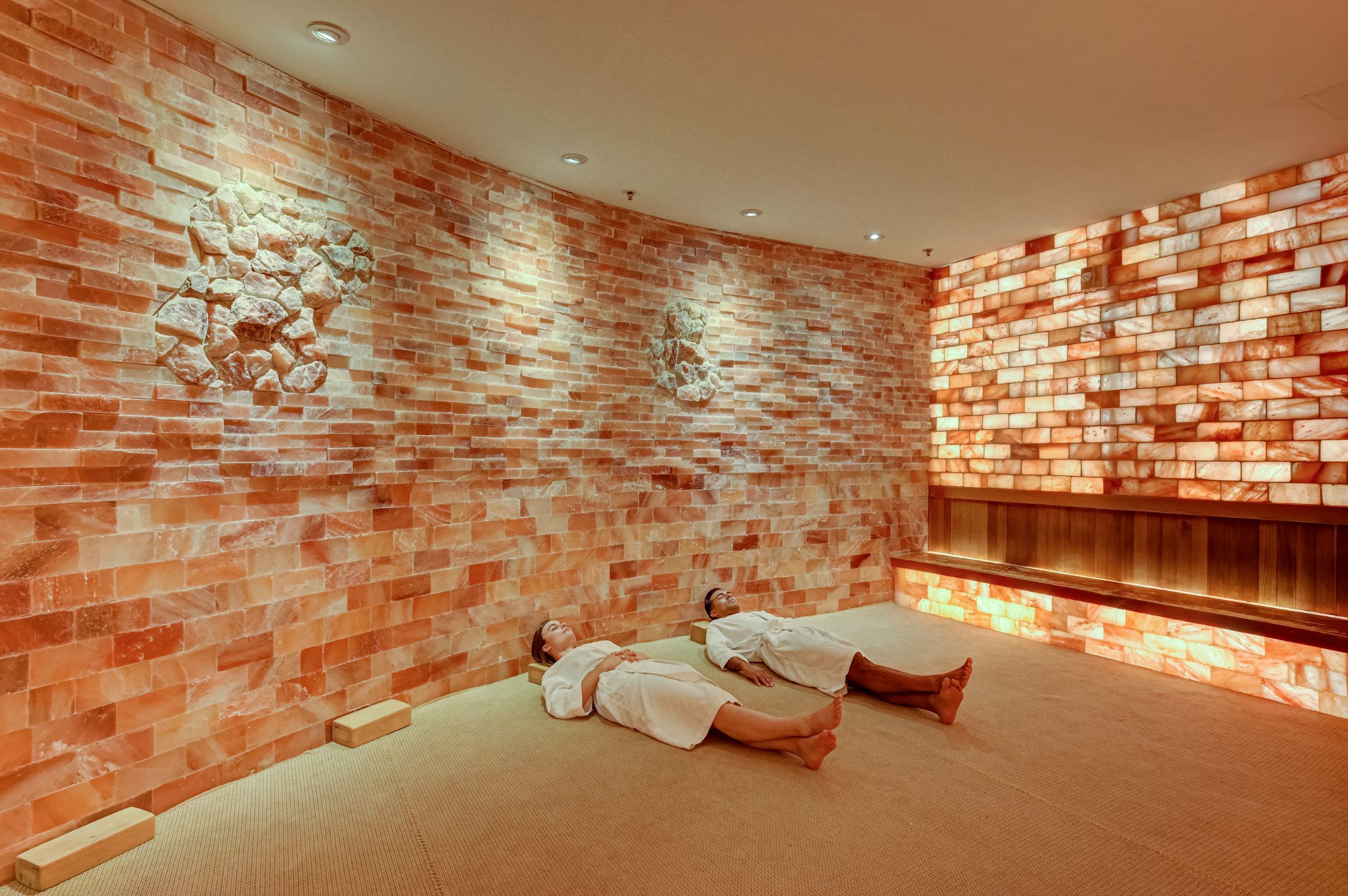 Two people lying on the ground of the Himalayan salt sauna at SoJo Spa Club, wearing robes, eyes closed, and relaxing