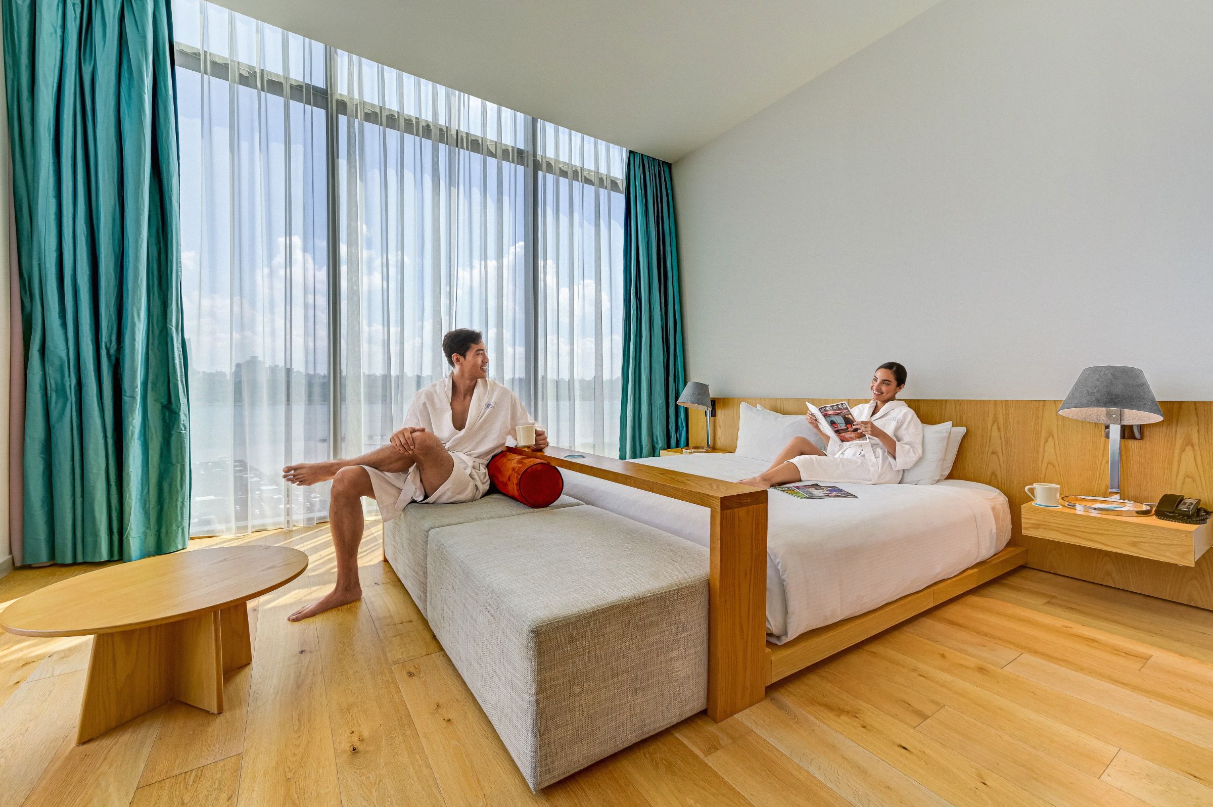 Couple enjoys coffee and reading in corner suite at Hotel Sojo. Man sits on ottoman while woman lies on king bed. Floor-to-ceiling windows showcase NYC view.