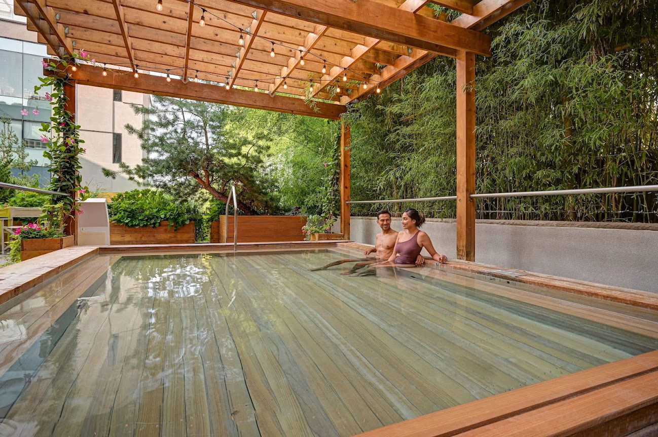 Couple sits in the hinoki bath surrounded by lush greenery at SoJo Spa Club.