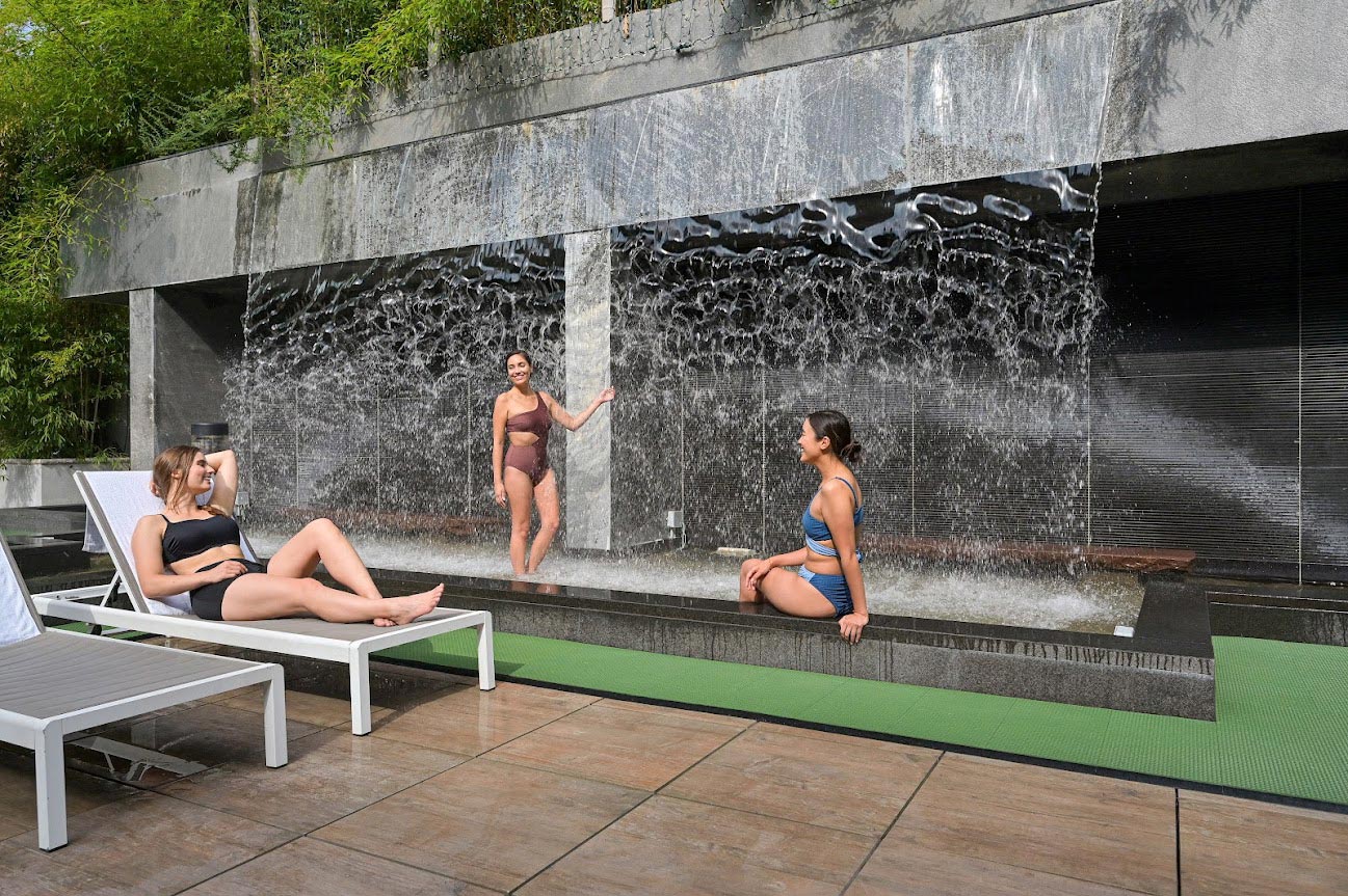 Three friends enjoying the waterfall feature at SoJo Spa Club. One walking through cold water, another lounging nearby, third sitting with feet in pool. Surrounded by greenery, with tranquil sound of waterfall.