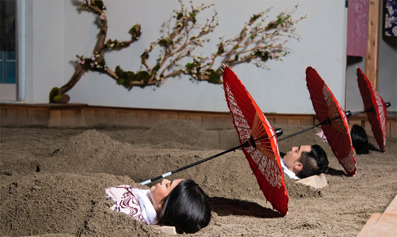 Three people laying in sand with their heads resting on a wooden block. Each with a red umbrella laying above their head.