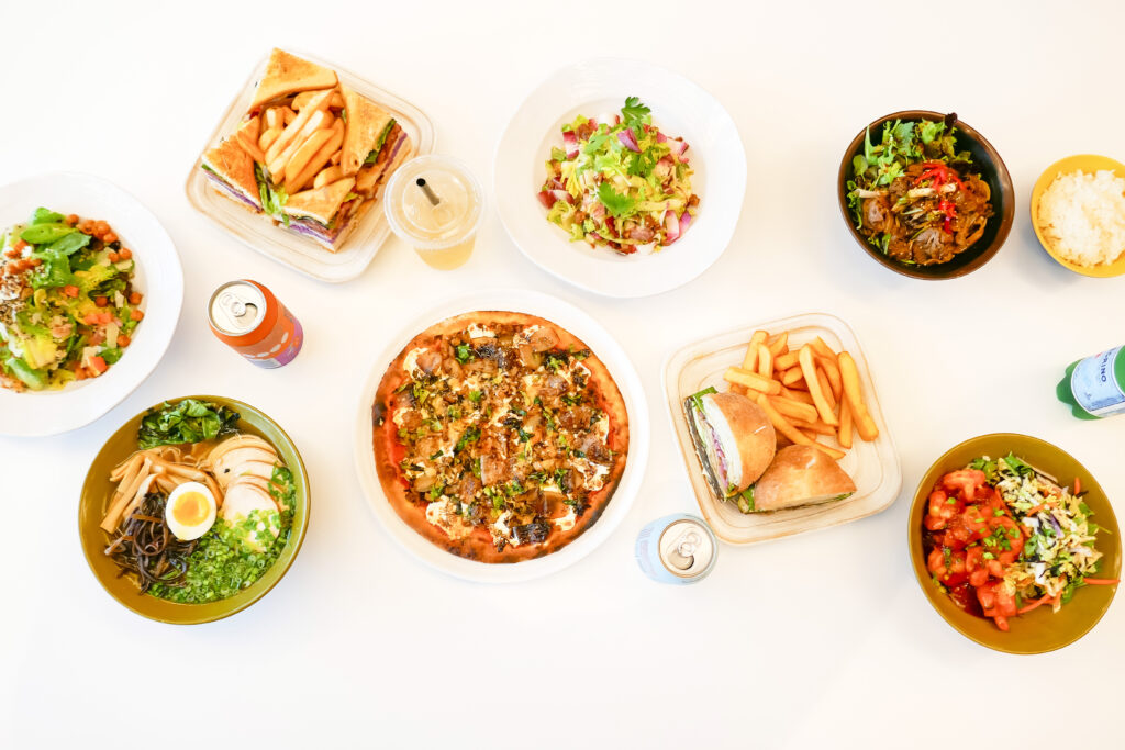 From Left to Right the Harvest Quinoa Salad, Chicken Club, Chidken Ramen, Sausage PIzza, Autumn Chopped Salad, Shrimp Bowl, Veggie Burger and Bulogi Bowl on a white table.