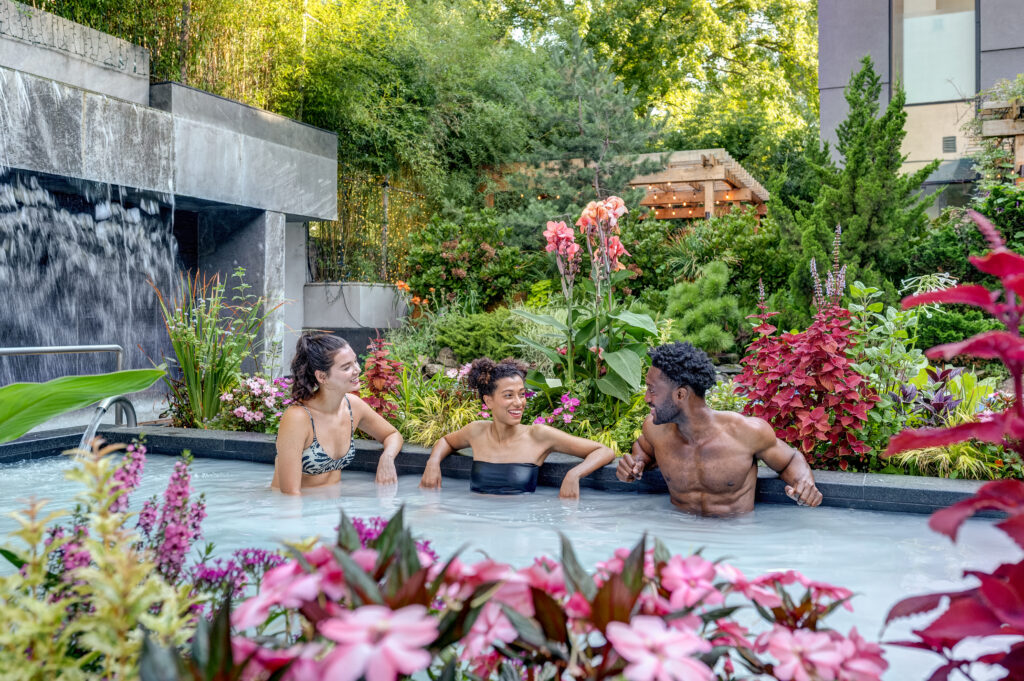 Two women and a man sit in the silk bath at SoJo Spa Club, surrounded by vibrant flowers, laughing and enjoying each ohter's company