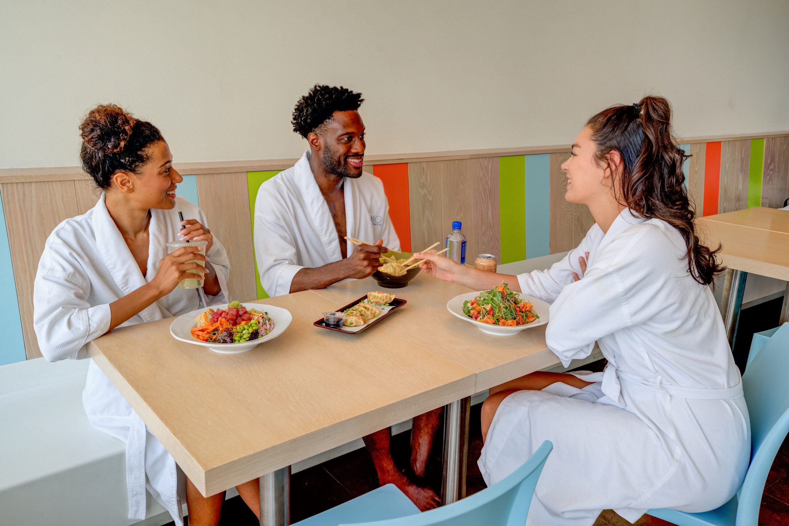 Two women and a man laugh while enjoying a lunch consisting of a Tuna Poke Bowl, Dumplings, Shirmp & Soba Noodle Salad and Chicken Ramen