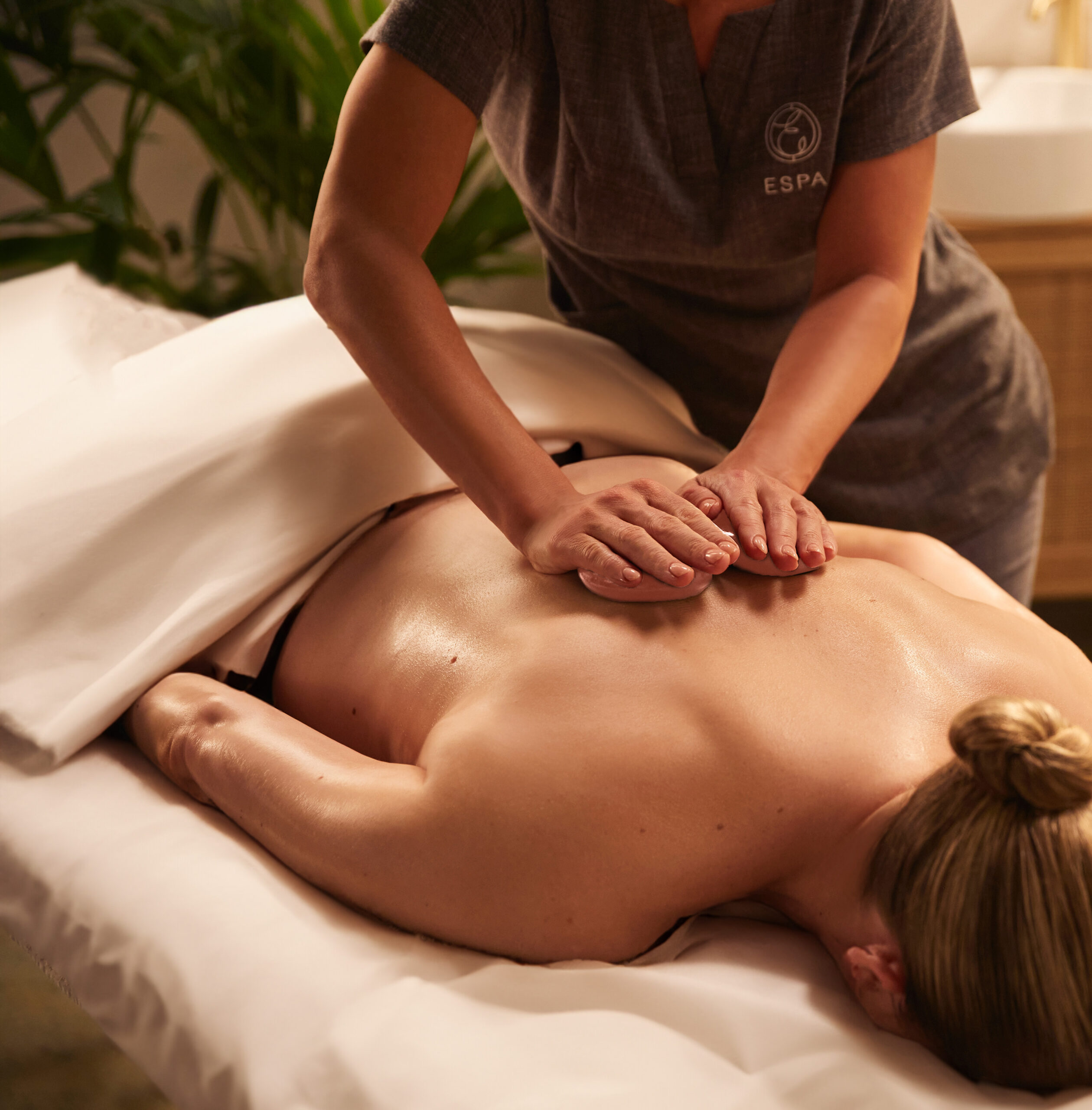 A massage therapist is performing a Heated Salt Stone massage on a client's back at SoJo Spa Club. The therapist's hands are gently pressing on the client's upper back with Himalayan Salt Stones.