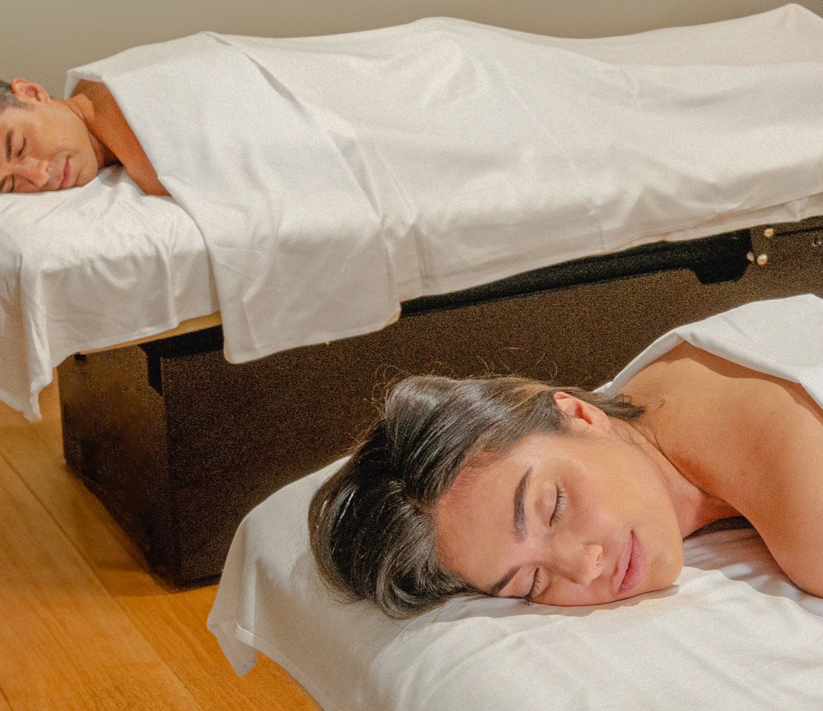 Two people relax side by side in a tranquil spa room, lying on massage tables with soft white linens, preparing for a couples massage.