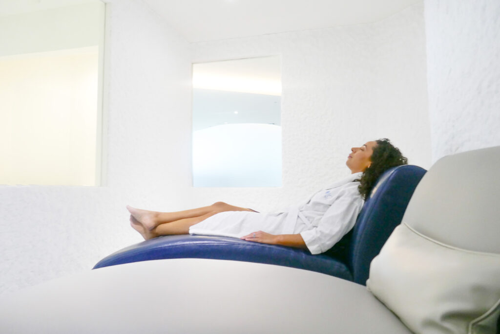 Woman relaxing on a blue lounge chair in a white room, enjoying the Halotherapy treatment for allergy relief.