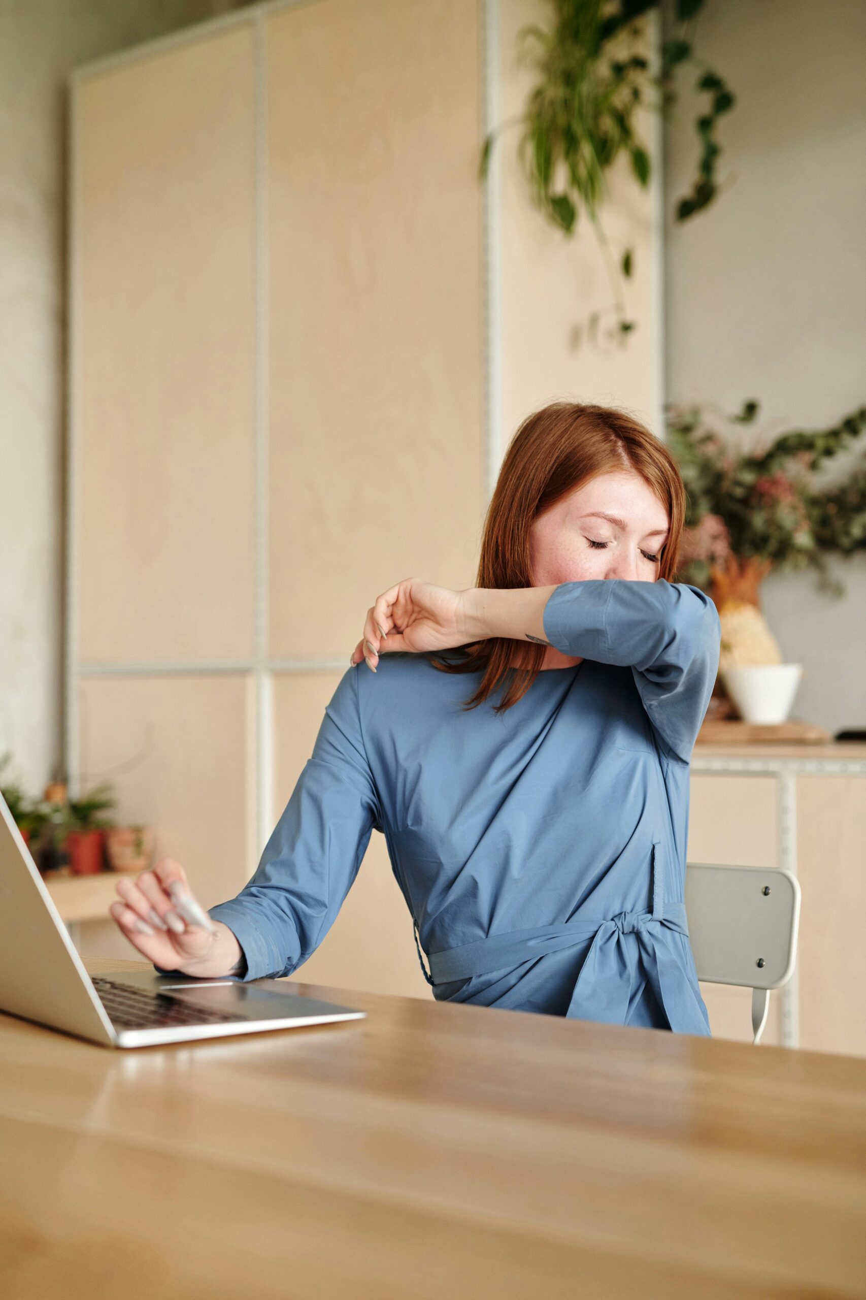 Woman wearing a blue shirt and sitting at her computer at a desk, sneezing into her elbow, during allergy season.